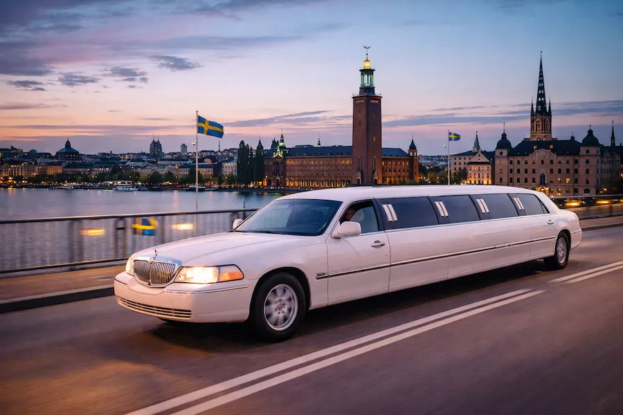 Limousine som kör genom Stockholm i stadsmiljö – vit stretchbil passerar över bro med Stadshuset och Riddarholmskyrkan i bakgrunden vid solnedgång.